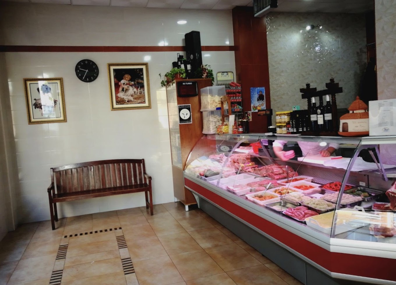 Interior de una carnicería tradicional con mostrador de carnes y embutidos, una banca de madera, cuadros decorativos en la pared y productos gourmet en estanterías.