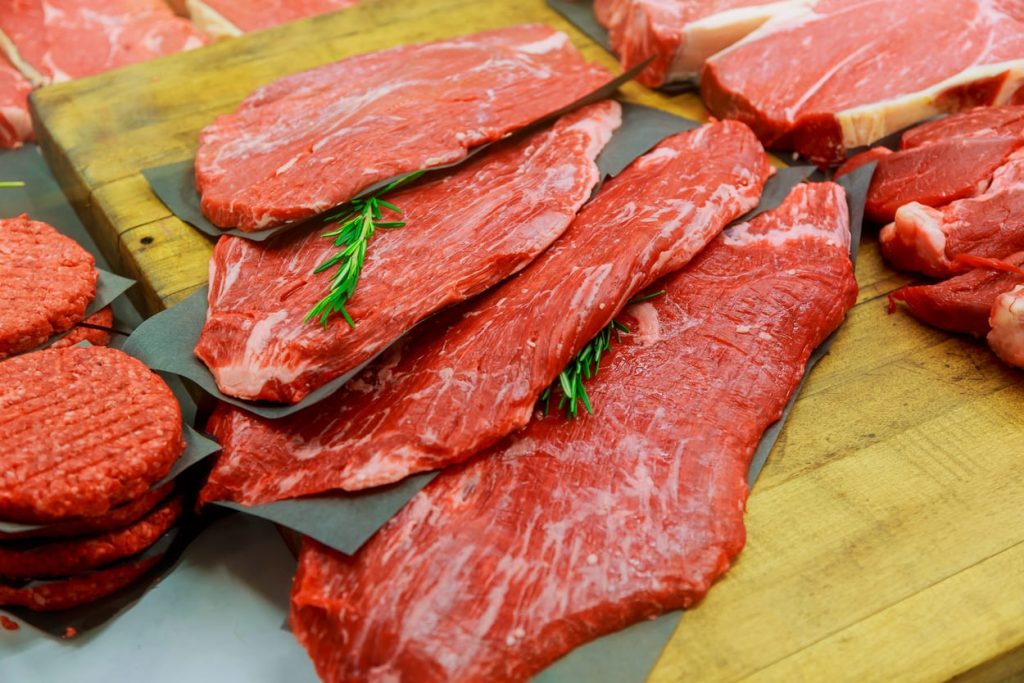 Varias piezas de carne roja y hamburguesas crudas sobre papel encerado en una tabla de cortar de madera, adornadas con ramitas de romero.