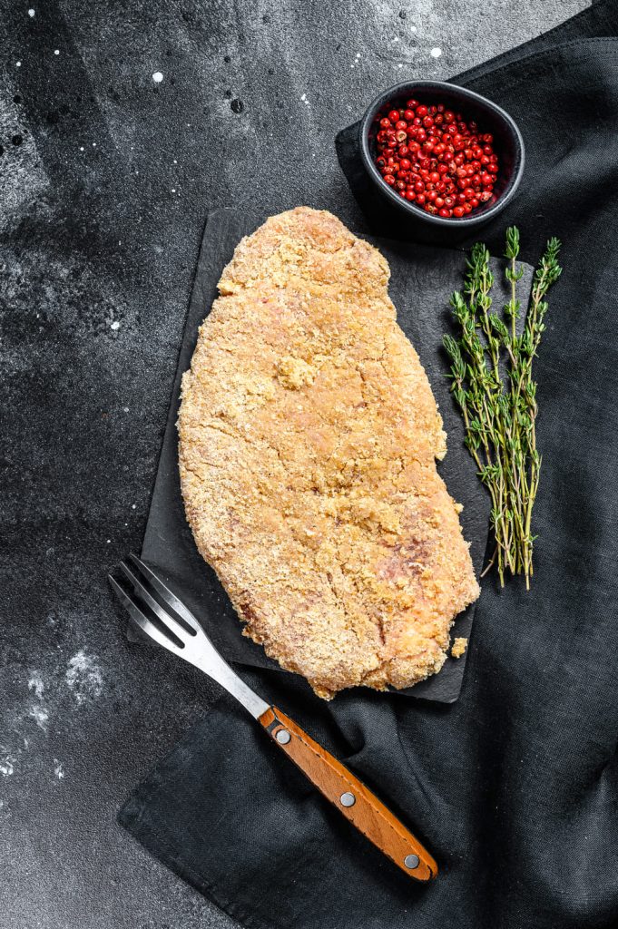 Fotografía de un filete de pescado empanizado con especias y hierbas en una tabla de cortar de madera sobre un mantel de lino, con una ramita de tomillo fresco encima y una mezcla de pimienta en un cuenco a un lado.